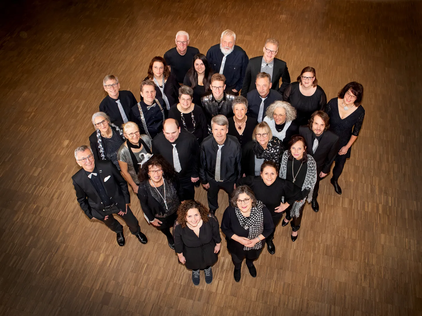 Der Chor von oben fotografiert: rund dreißig Sängerinnen und Sänger in Schwarz mit silbernen Krawatten und Tüchern stehen dicht beieinander und schauen lachend nach oben.